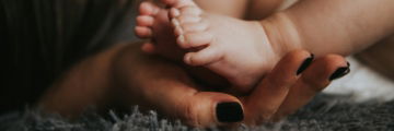 Infant's feet being held by a woman's hand with painted and manicured hands resting on a gray blanket